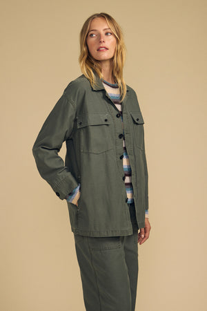 A woman stands against a beige background wearing the KAY SHIRT JACKET by Velvet by Graham & Spencer and matching pants over a striped sweater, with one hand in her pocket.