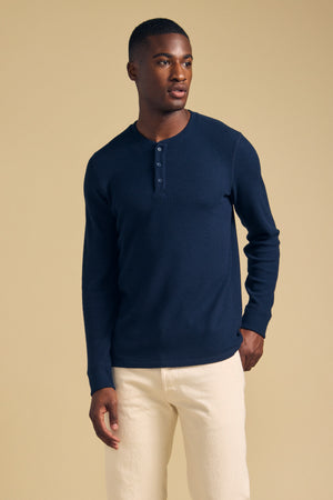 A man wearing a navy long-sleeved brushed thermal ROEN THERMAL HENLEY by Velvet by Graham & Spencer and cream-colored pants.