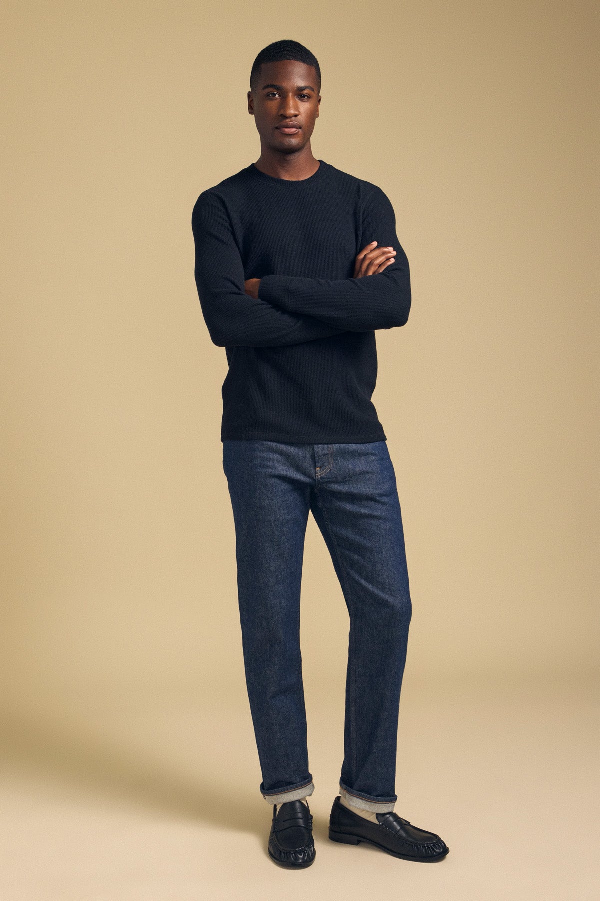 A man with arms crossed wearing a black Velvet by Graham & Spencer TOLLIVER THERMAL CREW PULLOVER, cuffed dark blue jeans, and black loafers.