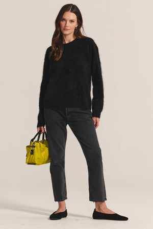 A woman stands against a plain background wearing the JACQUELINE CASHMERE SWEATER by Velvet by Graham & Spencer, black jeans, black flats, and holding a small yellow handbag.