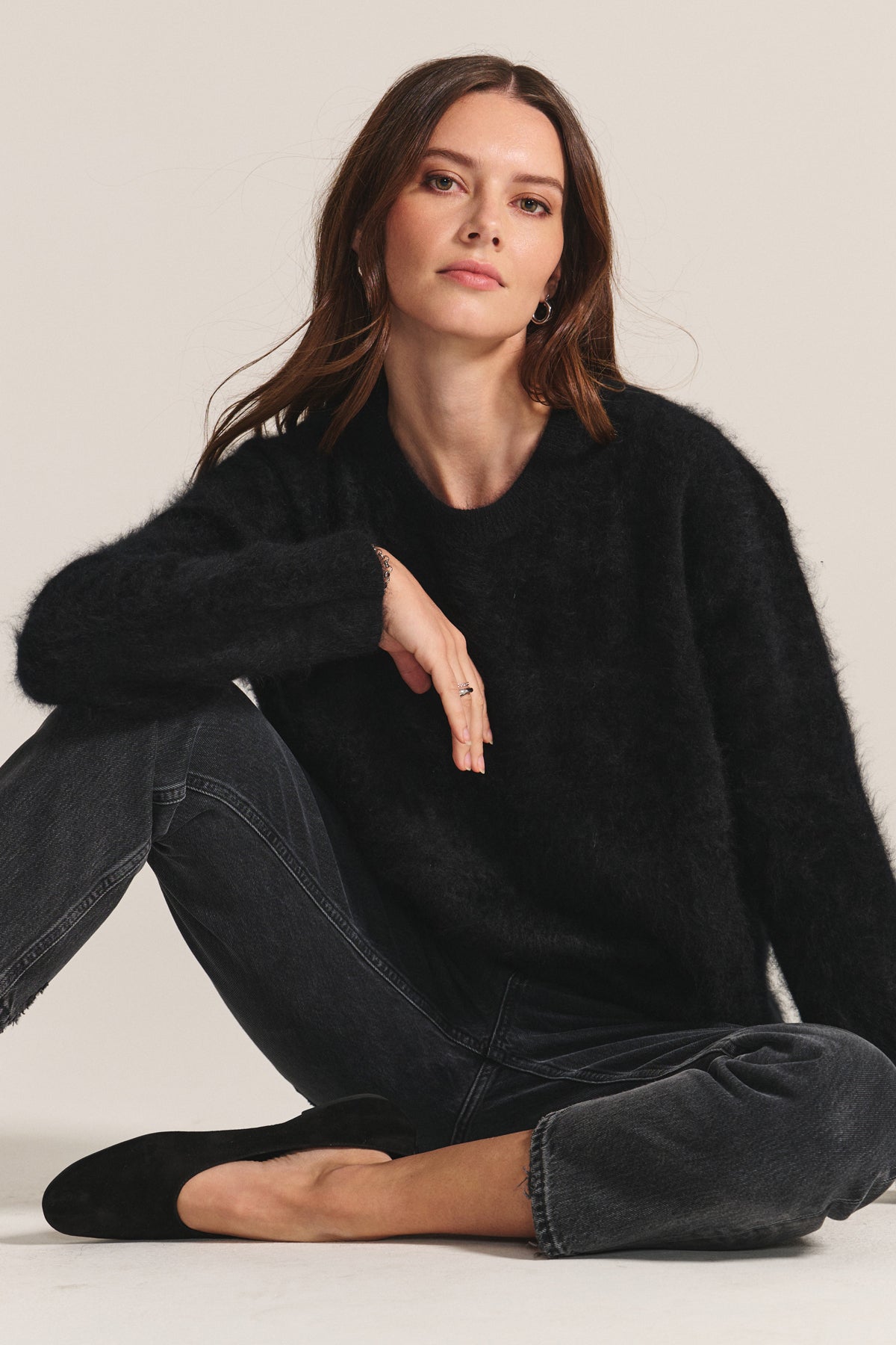 Woman with long brown hair wearing the Velvet by Graham & Spencer JACQUELINE CASHMERE SWEATER and dark jeans sits cross-legged on the floor against a plain background.