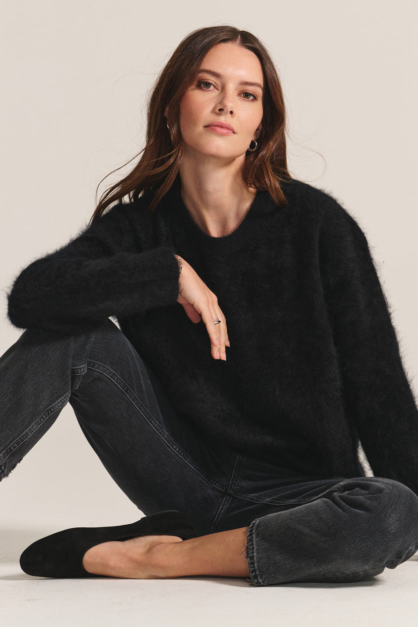 Woman with long brown hair wearing the Velvet by Graham & Spencer JACQUELINE CASHMERE SWEATER and dark jeans sits cross-legged on the floor against a plain background.