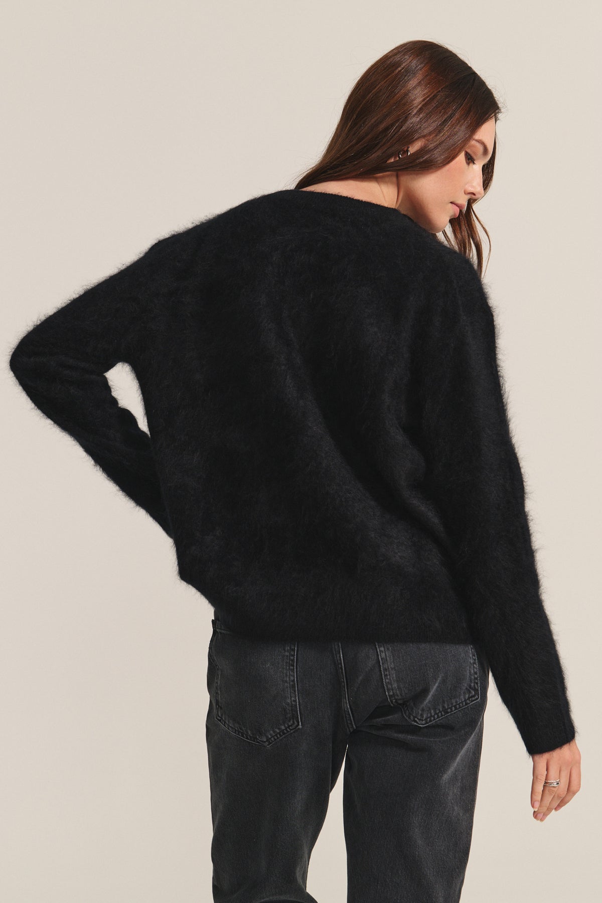 A person with long brown hair wears the JACQUELINE CASHMERE SWEATER by Velvet by Graham & Spencer and dark jeans, standing with their back turned and looking over their shoulder.