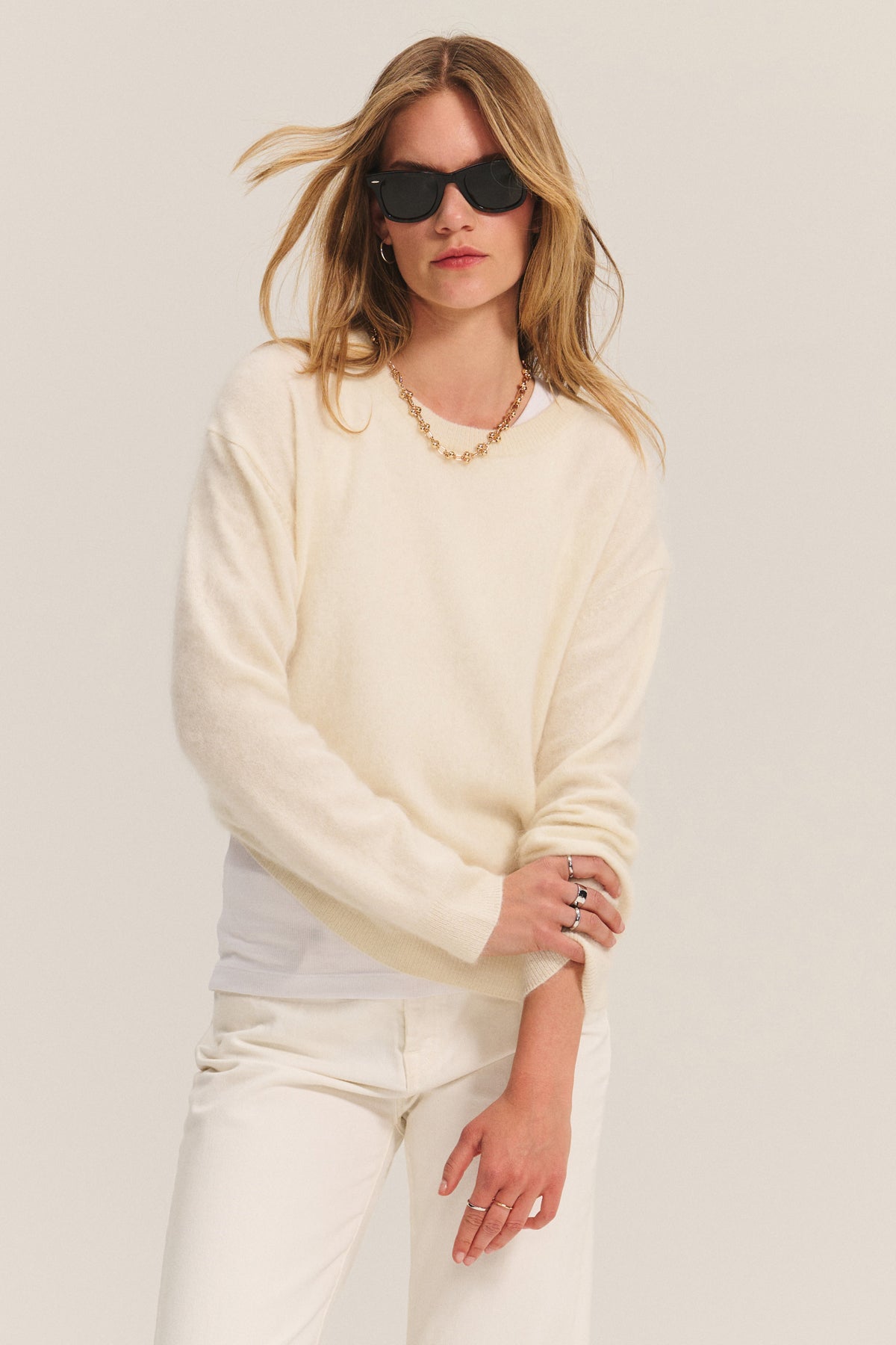 A woman wears black sunglasses, white pants, and the Velvet by Graham & Spencer JACQUELINE CASHMERE SWEATER in cream against a plain background.