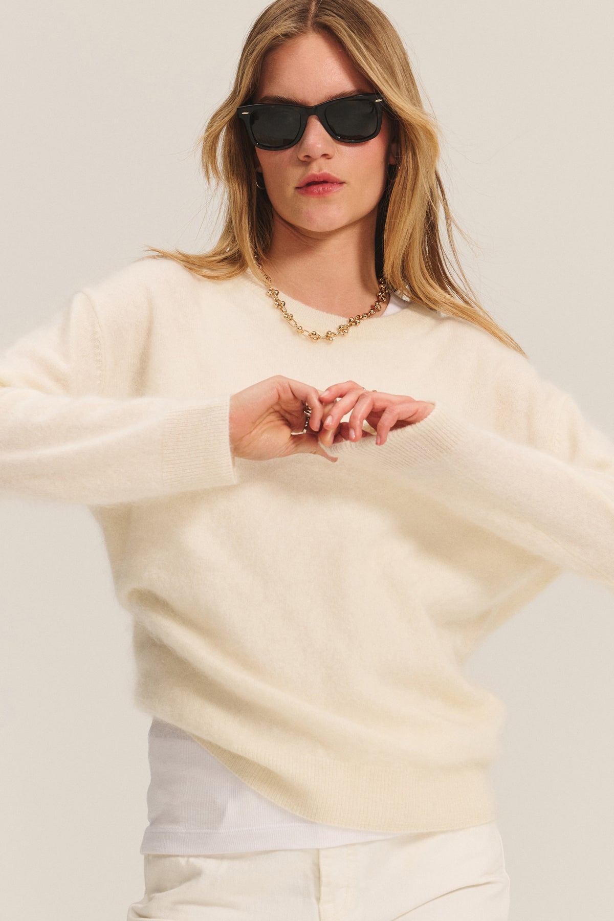 A woman stands against a plain background wearing black sunglasses, a gold chain necklace, and the JACQUELINE CASHMERE SWEATER in off-white by Velvet by Graham & Spencer over a white shirt.