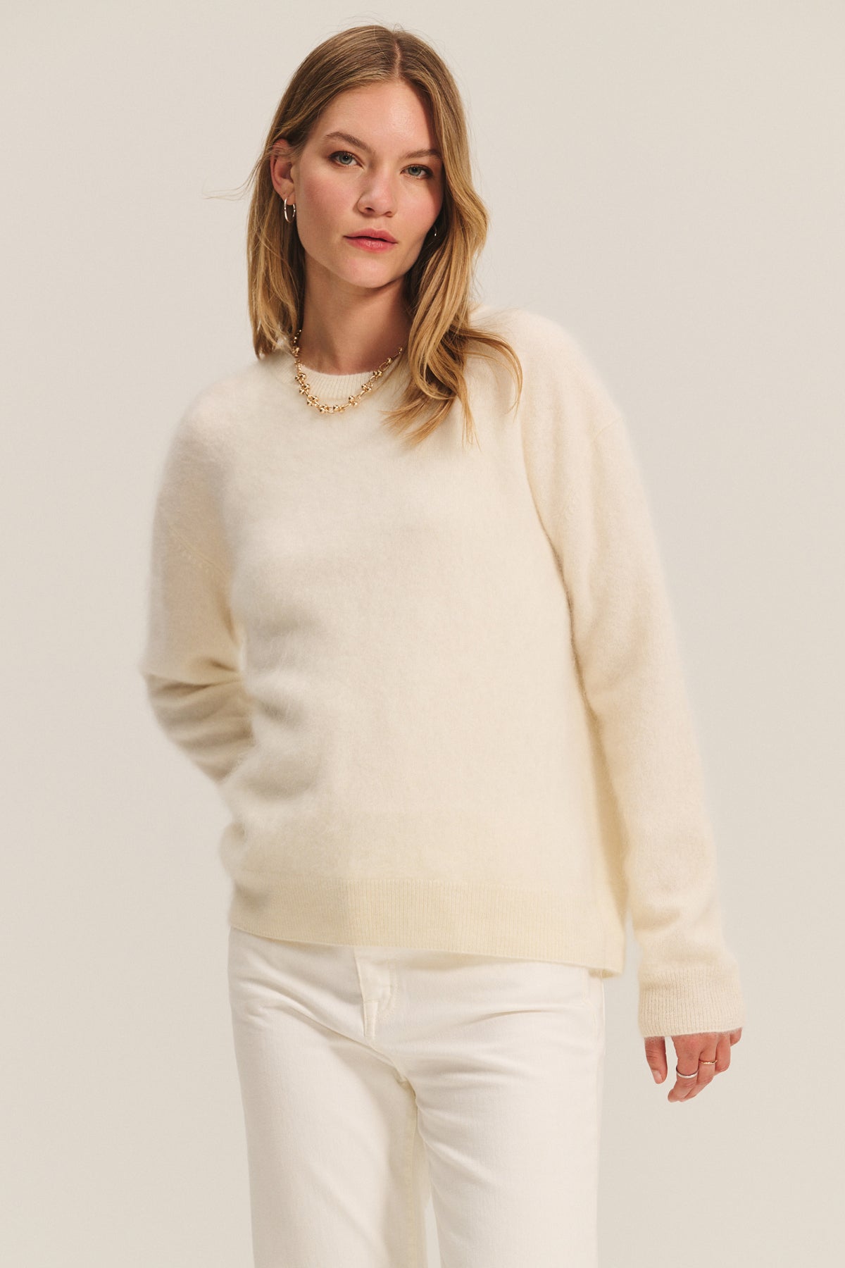 A woman wears the Velvet by Graham & Spencer JACQUELINE CASHMERE SWEATER in a luxurious cream shade with white pants, standing against a plain light background.