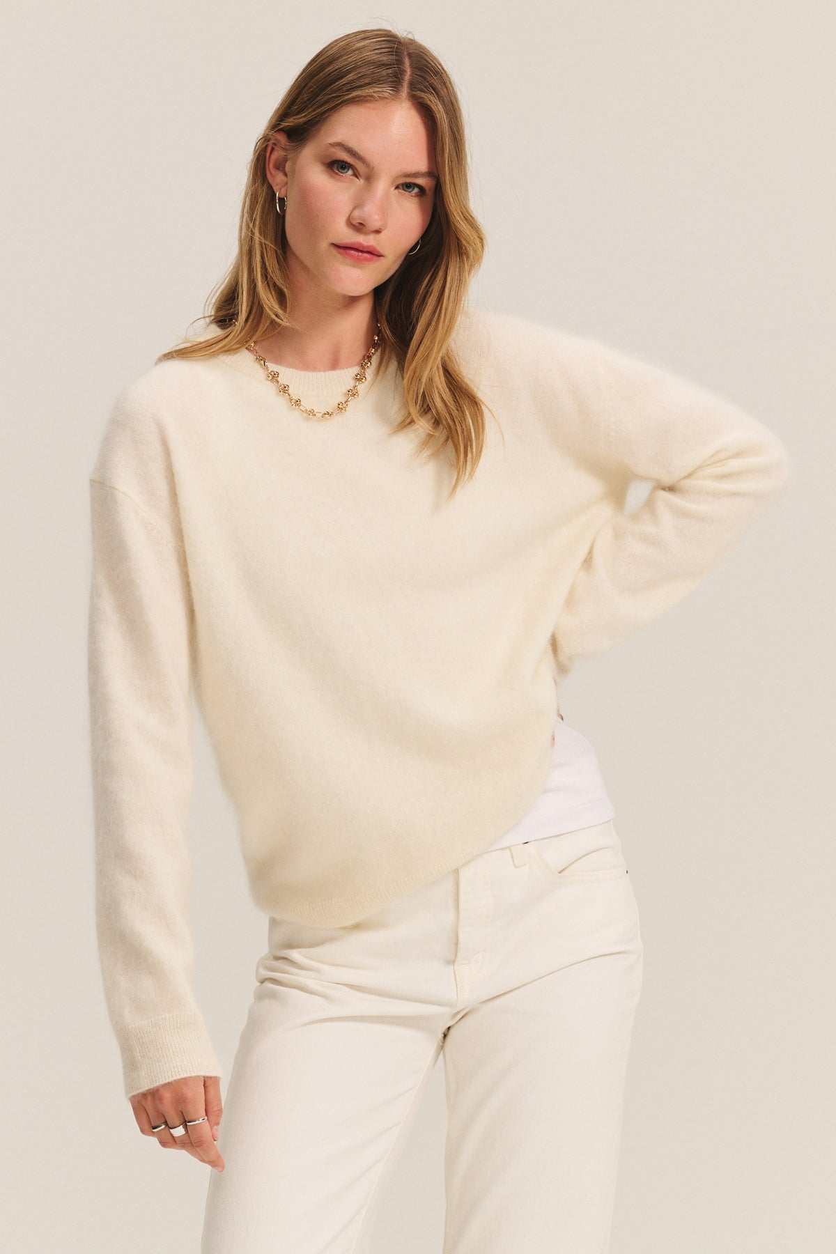 A woman wears the JACQUELINE CASHMERE SWEATER by Velvet by Graham & Spencer in cream, paired with white pants and a gold necklace, standing against a plain light background.