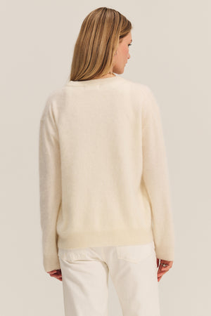 Seen from behind, a person with long light brown hair wears the JACQUELINE CASHMERE SWEATER by Velvet by Graham & Spencer in off-white, paired with white pants against a plain background.