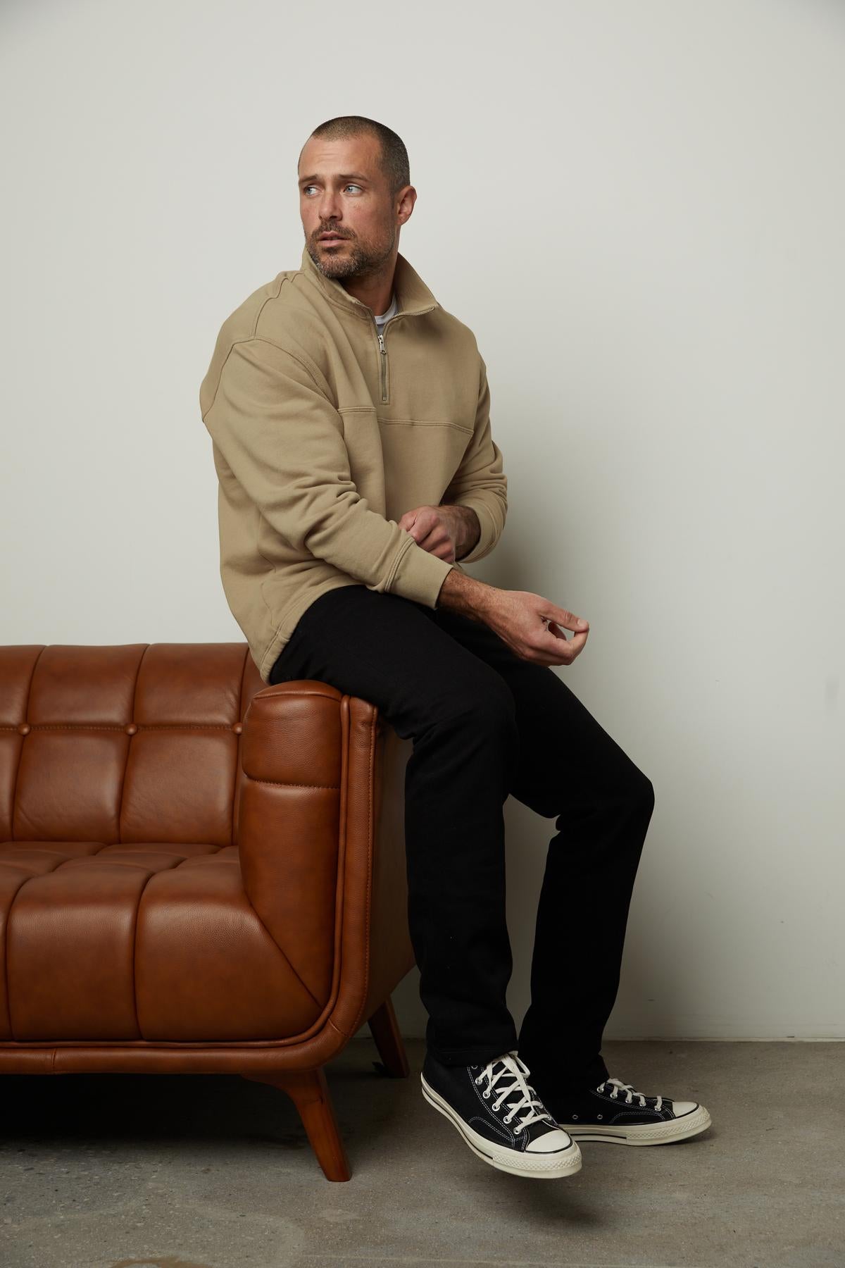   A man sitting on a brown leather couch, wearing a BOSCO QUARTER-ZIP SWEATSHIRT by Velvet by Graham & Spencer. 
