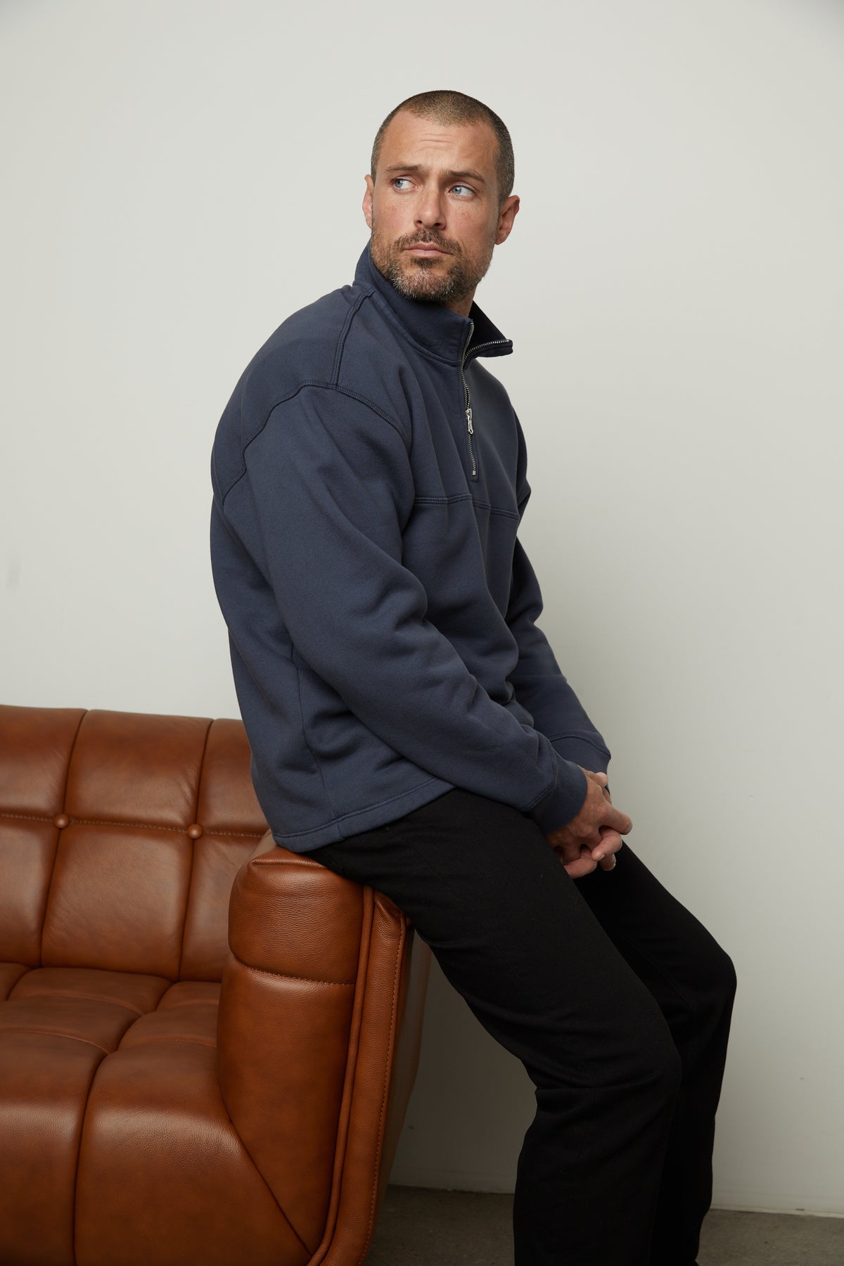 A man sitting on a brown leather couch, wearing a Velvet by Graham & Spencer BOSCO QUARTER-ZIP SWEATSHIRT.-35782782714049