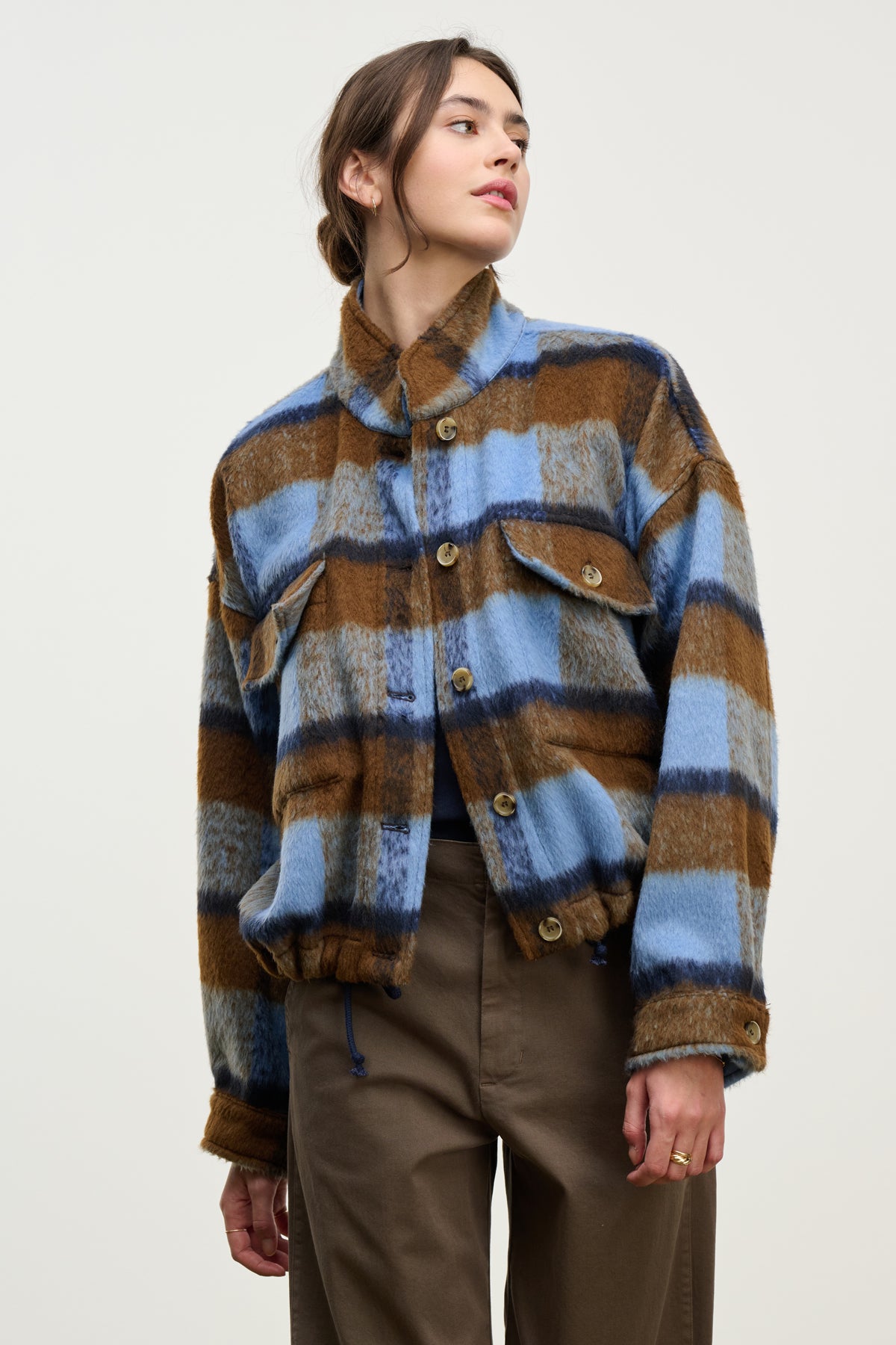 A person wears the AUBREY JACKET by Velvet by Graham & Spencer—a blue, brown, and gray plaid piece featuring a stand-up collar and flap pockets—while standing against a plain light background.-39814813548737