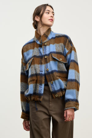 A person wears the AUBREY JACKET by Velvet by Graham & Spencer—a blue, brown, and gray plaid piece featuring a stand-up collar and flap pockets—while standing against a plain light background.