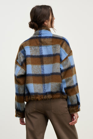 A person with brown hair in a low bun faces away from the camera, wearing the AUBREY JACKET by Velvet by Graham & Spencer in blue and brown plaid with an adjustable hem, paired with brown pants against a plain light background.