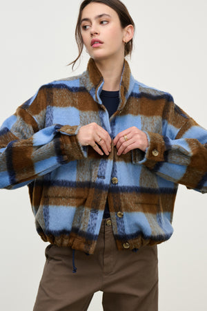 Woman wears the AUBREY JACKET by Velvet by Graham & Spencer—a blue and brown plaid style with stand-up collar and adjustable hem—standing with hands near buttons against a plain background.