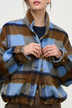 A woman wears the AUBREY JACKET by Velvet by Graham & Spencer—a blue, brown, and white plaid piece with a stand-up collar and adjustable hem. Her hands are near the buttons; her face is partially out of frame.