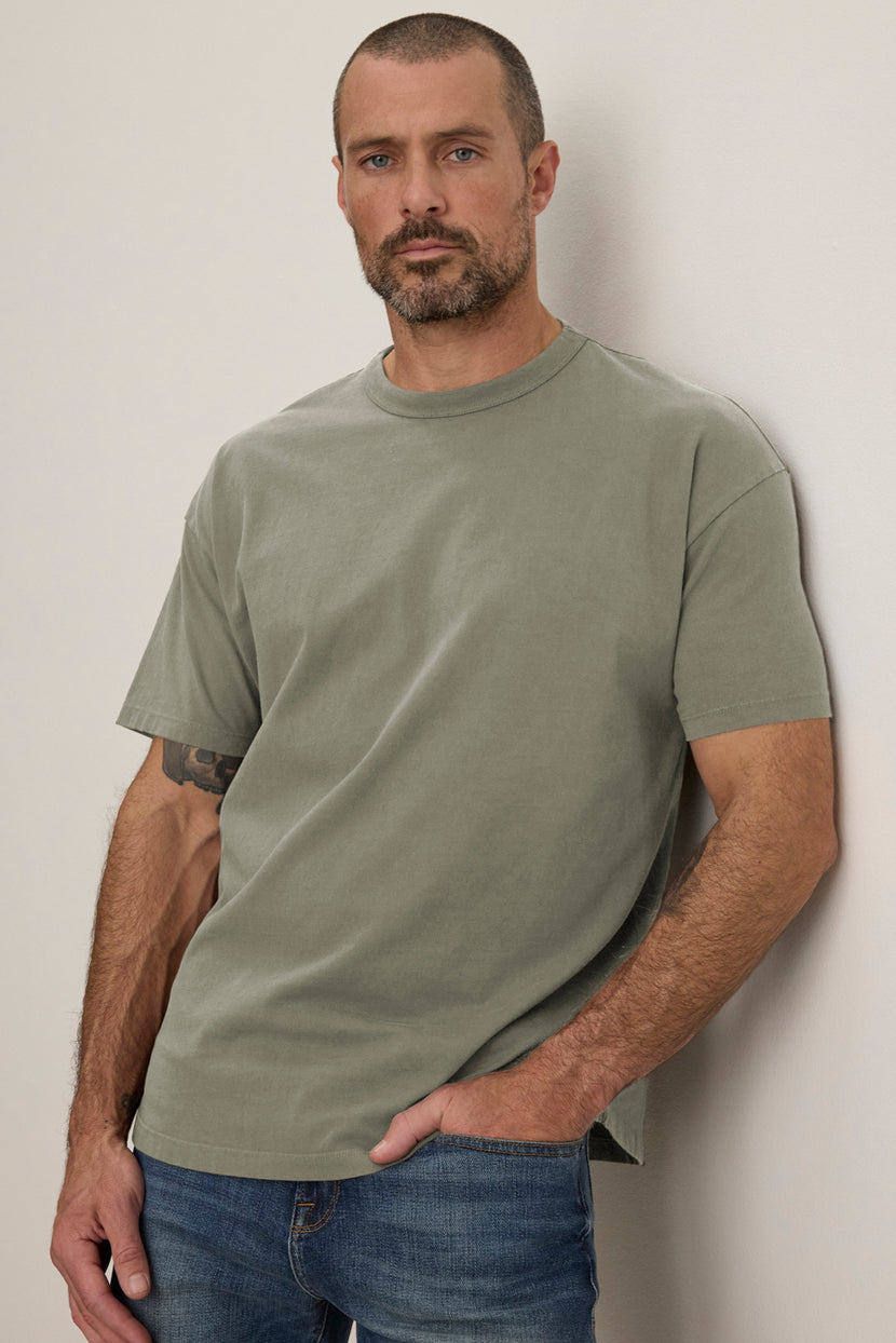 A man with short hair and a beard stands against a light-colored wall, wearing the BRYAN TEE by Velvet by Graham & Spencer, a gray, relaxed-fit t-shirt, along with blue jeans.