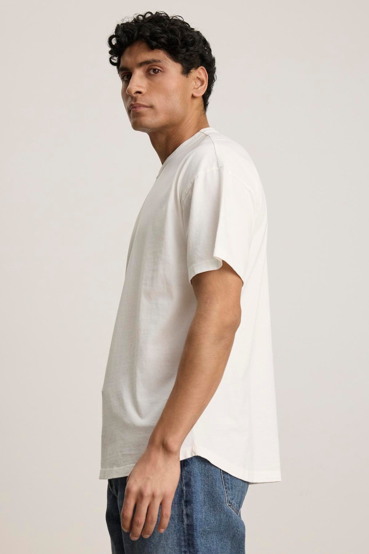   A person with curly hair wearing the BRYAN TEE by Velvet by Graham & Spencer made from pima cotton jersey and blue jeans stands sideways against a neutral background. 