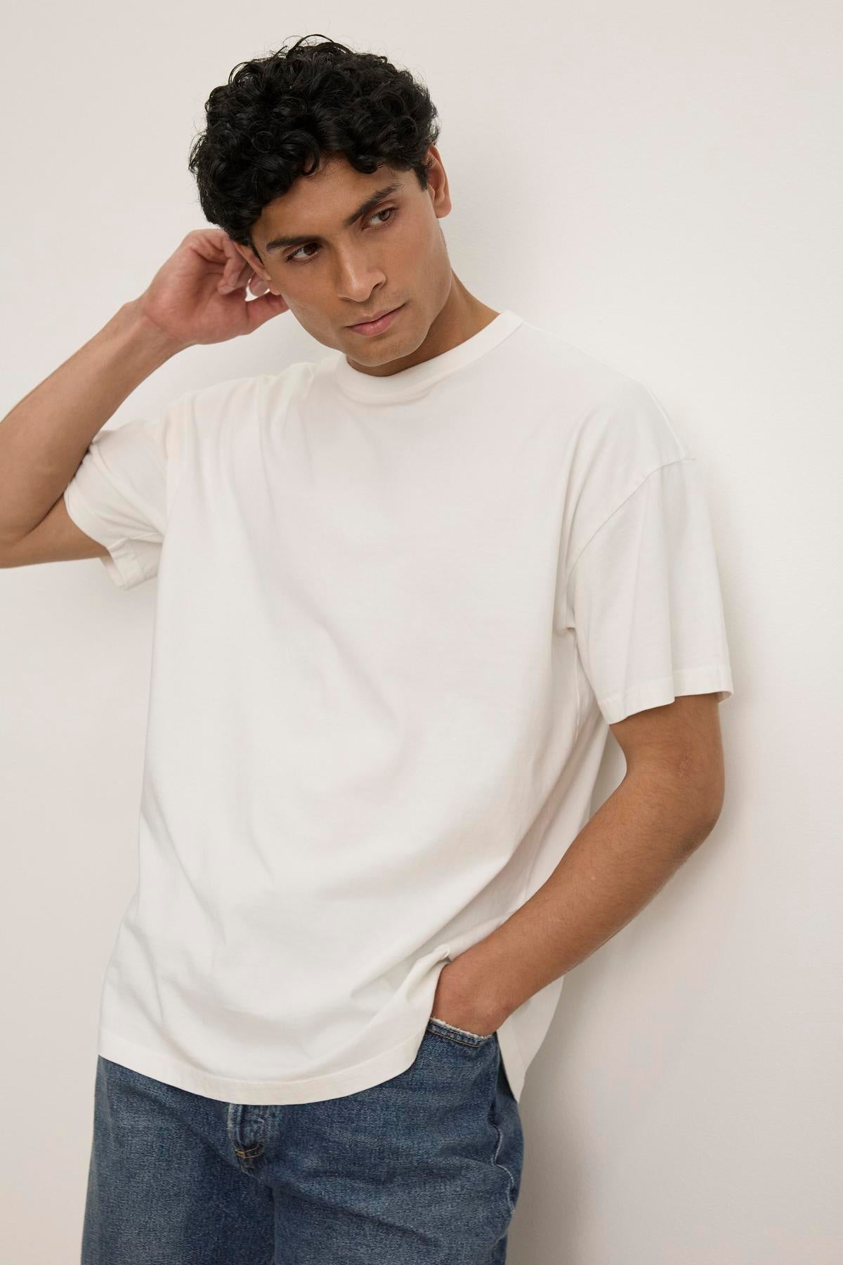   A person wearing a relaxed-fit BRYAN TEE by Velvet by Graham & Spencer and jeans stands against a plain white background, with one hand in a pocket and the other touching their neck. 