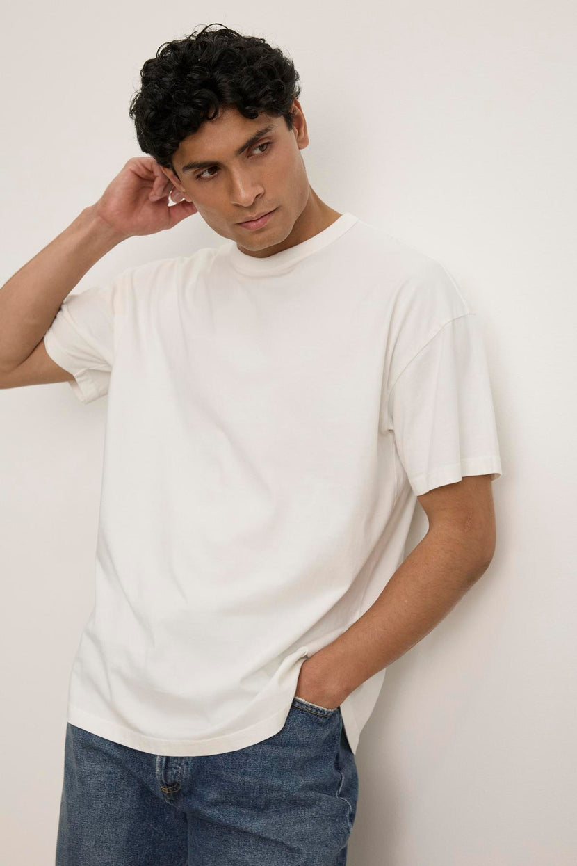 A person wearing a relaxed-fit BRYAN TEE by Velvet by Graham & Spencer and jeans stands against a plain white background, with one hand in a pocket and the other touching their neck.