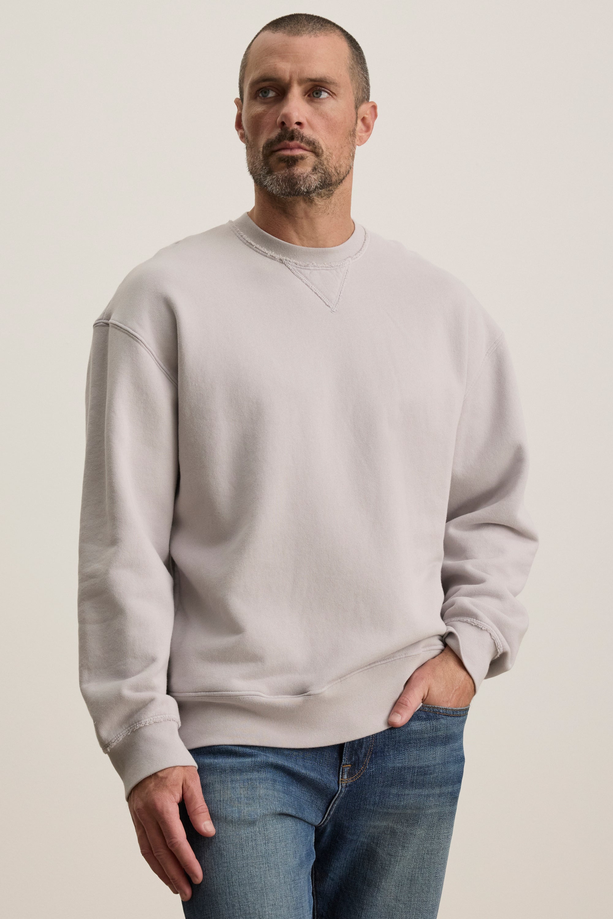   A man wearing a Velvet by Graham & Spencer FORREST SWEATSHIRT in relaxed-fit, light gray cotton fleece and blue jeans stands with one hand in his pocket against a plain background. 