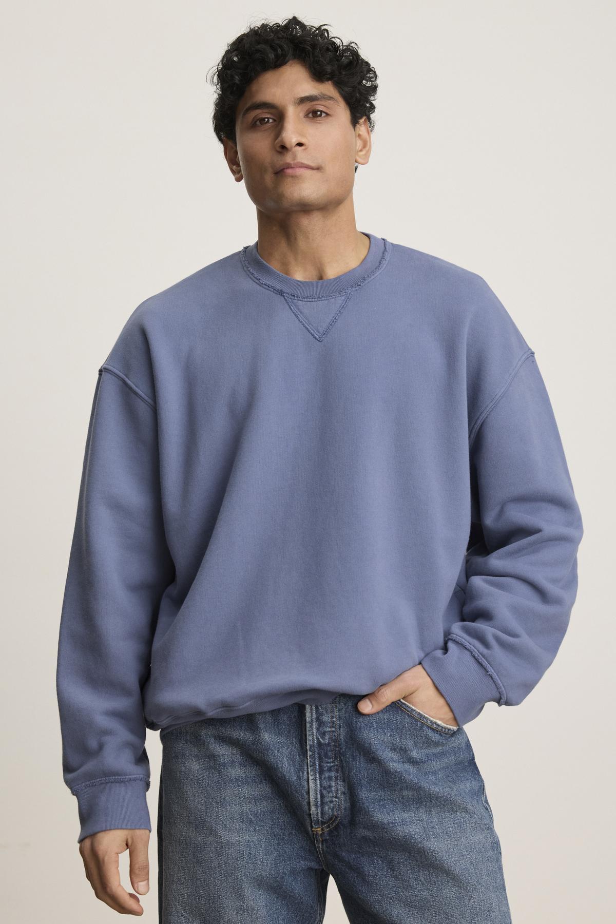   A person models the FORREST SWEATSHIRT, a relaxed fit piece by Velvet by Graham & Spencer, pairing it with jeans. They stand against a plain backdrop, highlighting the garment-dyed cotton fleece and raw edge details with one hand in their pocket. 