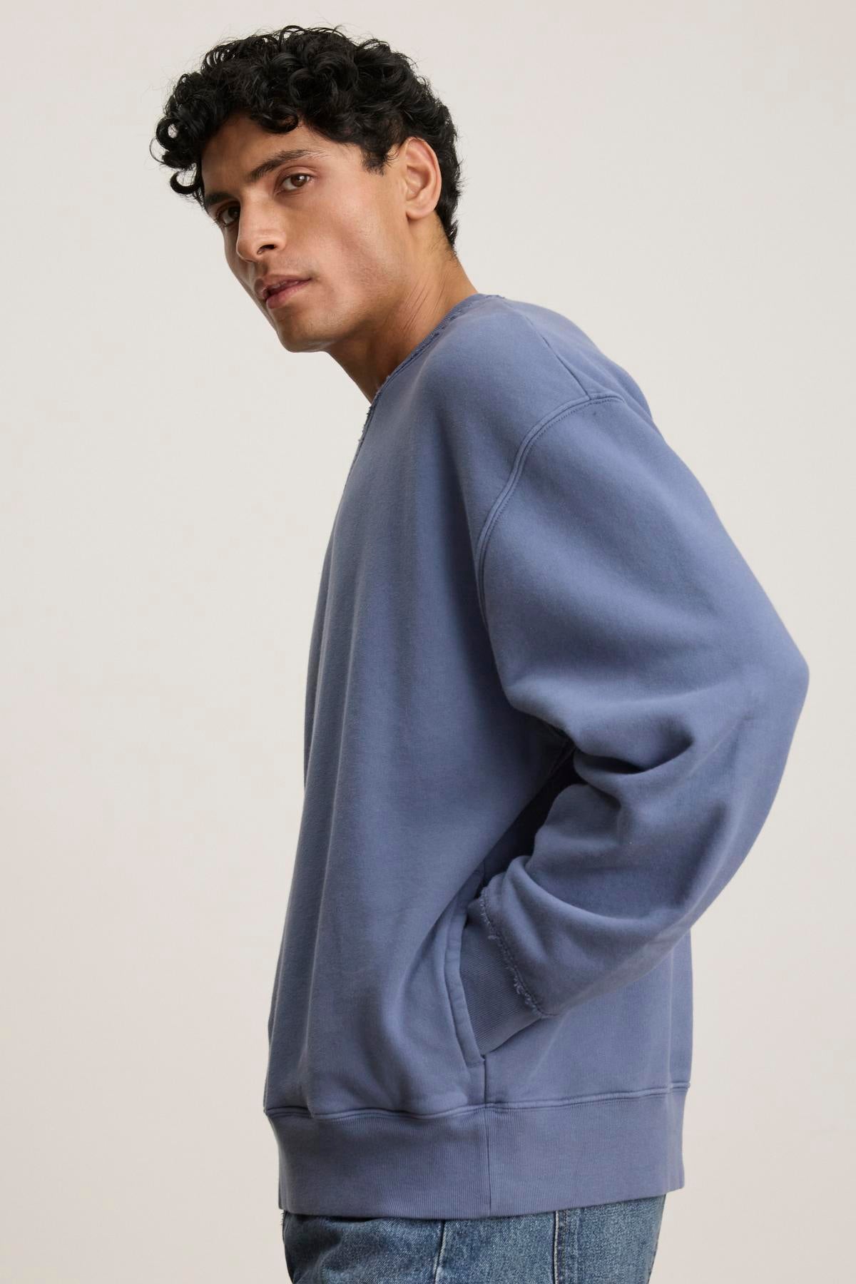 Wearing a blue FORREST SWEATSHIRT by Velvet by Graham & Spencer, with ribbed trims and crafted from heavyweight cotton fleece, a person stands with hands in pockets, looking sideways.-38778834714817