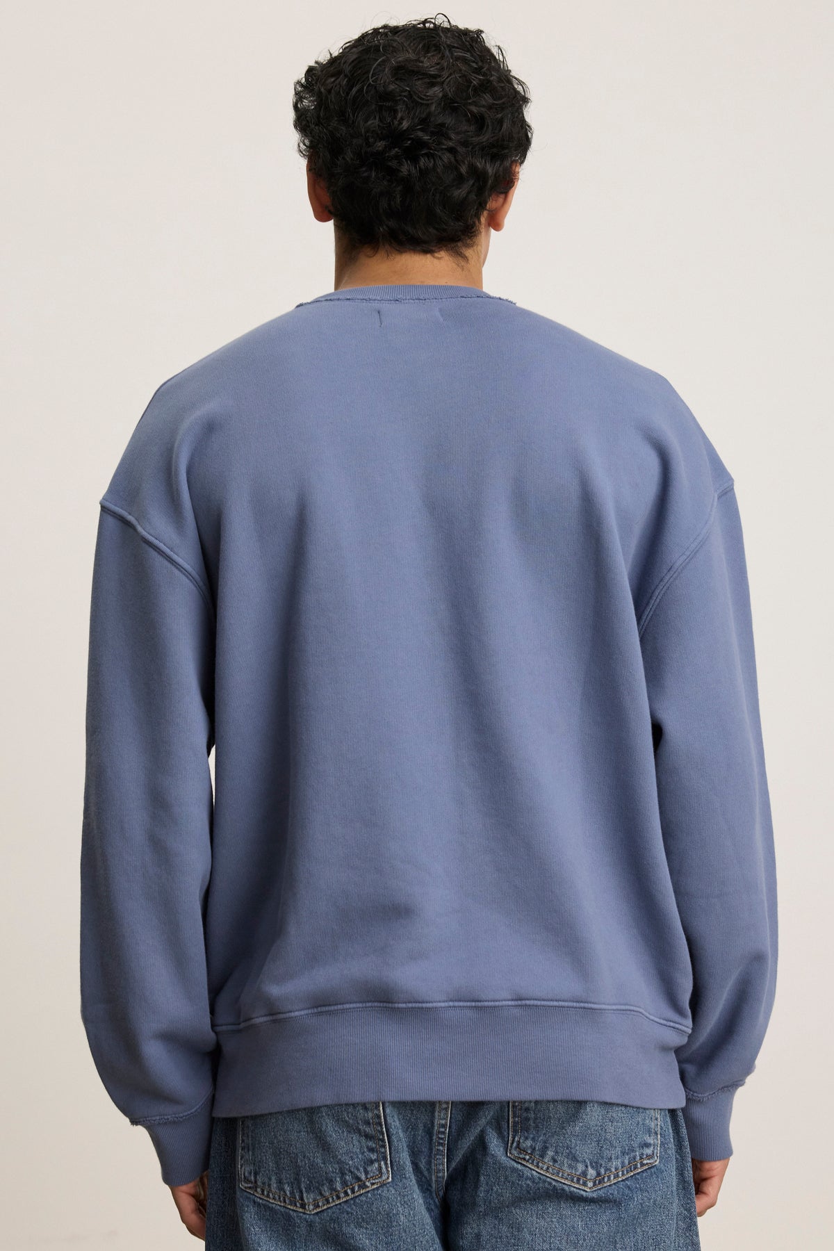   Someone with curly hair wears Velvet by Graham & Spencer's FORREST SWEATSHIRT, a blue heavyweight cotton fleece with ribbed trims and a relaxed fit, paired with jeans. They stand casually facing away. 