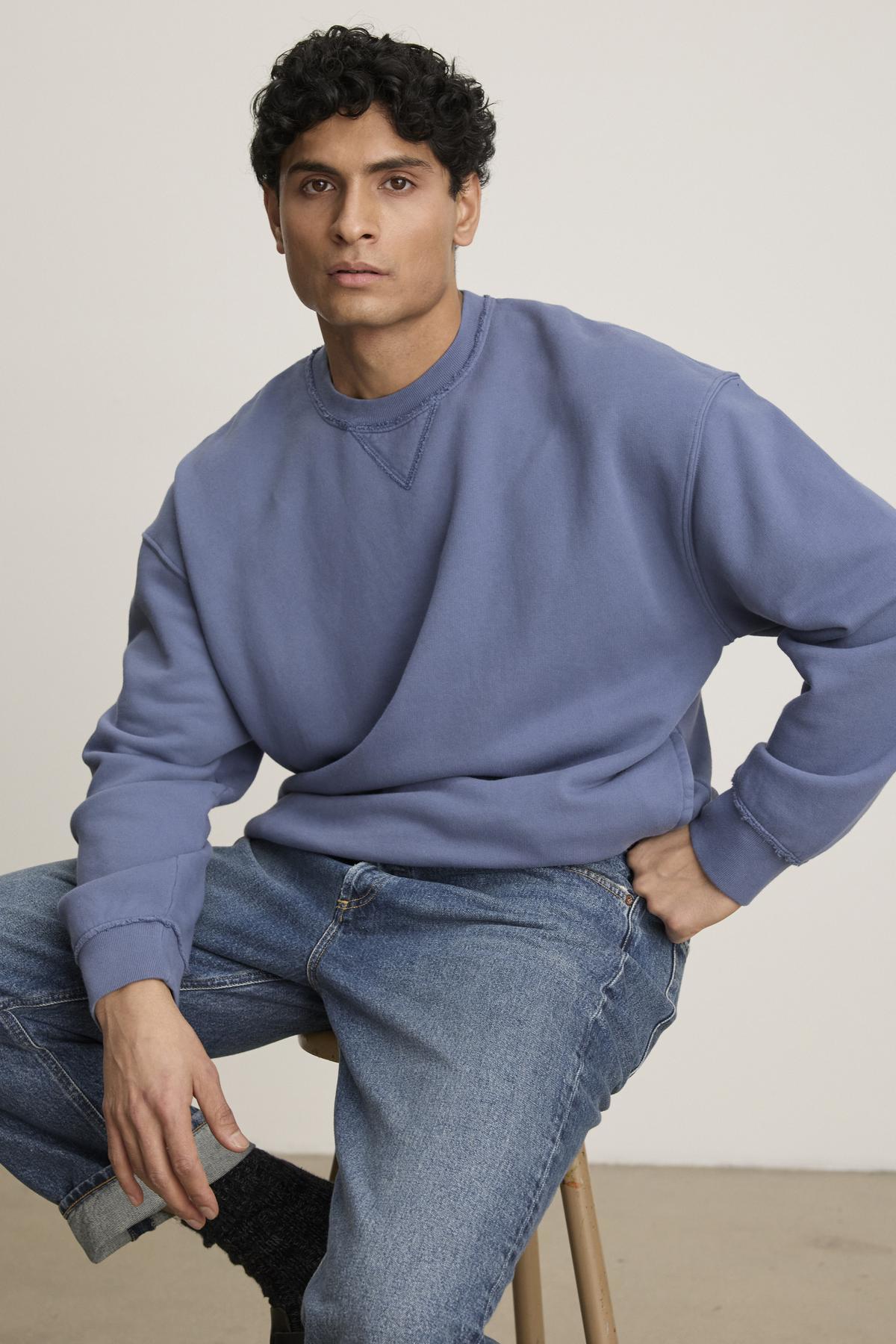   A person lounges in Velvet by Graham & Spencer's FORREST SWEATSHIRT—a blue heavyweight cotton fleece with ribbed trims and relaxed fit—paired with jeans while sitting on a stool against a plain backdrop, exuding casual comfort. 