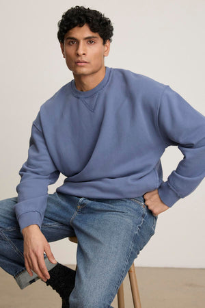 A person lounges in Velvet by Graham & Spencer's FORREST SWEATSHIRT—a blue heavyweight cotton fleece with ribbed trims and relaxed fit—paired with jeans while sitting on a stool against a plain backdrop, exuding casual comfort.