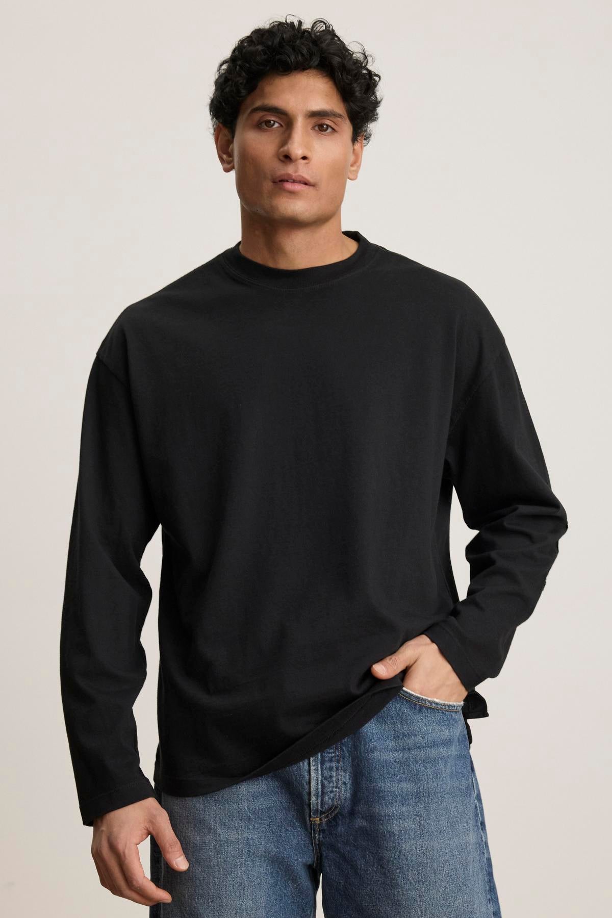   A person stands in a Velvet by Graham & Spencer SLATER TEE, embodying a relaxed fit that easily complements any style against the plain background. 