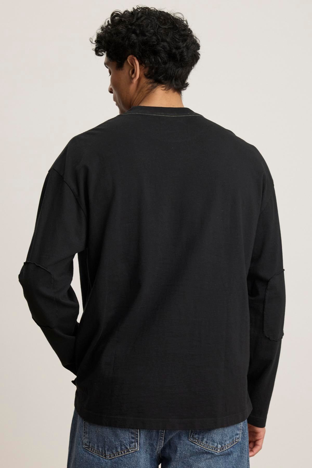   A person with curly hair is wearing a SLATER TEE by Velvet by Graham & Spencer, featuring a black long-sleeve relaxed fit, along with blue jeans, facing away from the camera. 