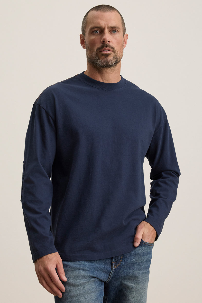 A short-haired, bearded individual stands against a neutral background wearing Velvet by Graham & Spencer's SLATER TEE, a dark blue long-sleeve pima cotton jersey, paired with jeans. It offers a relaxed fit suitable for any occasion.