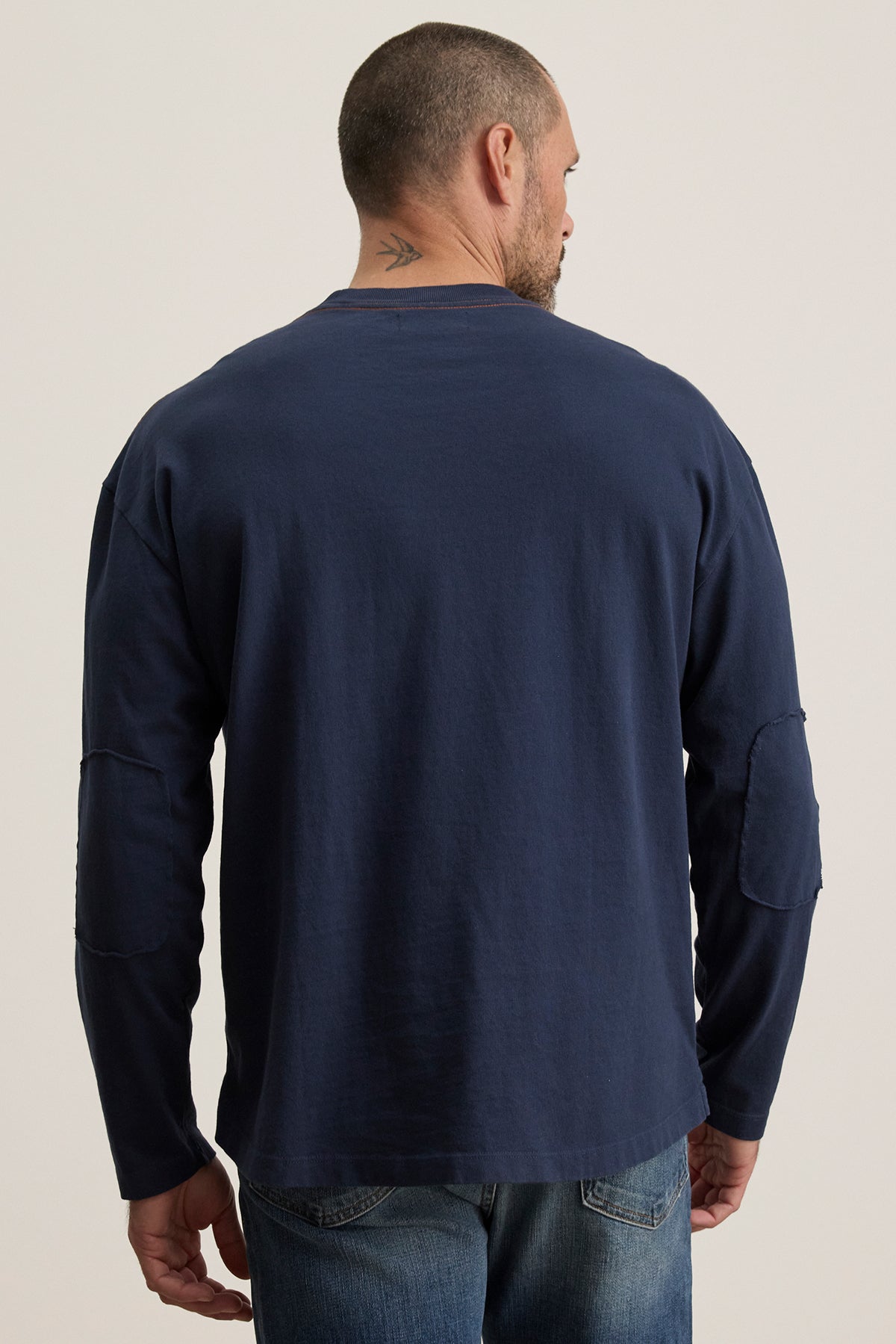 A man stands with his back turned, wearing Velvet by Graham & Spencer's SLATER TEE in navy, paired with blue jeans. The relaxed fit ensures comfort, and a tattoo on the back of his neck adds an element of intrigue.-38500088709313