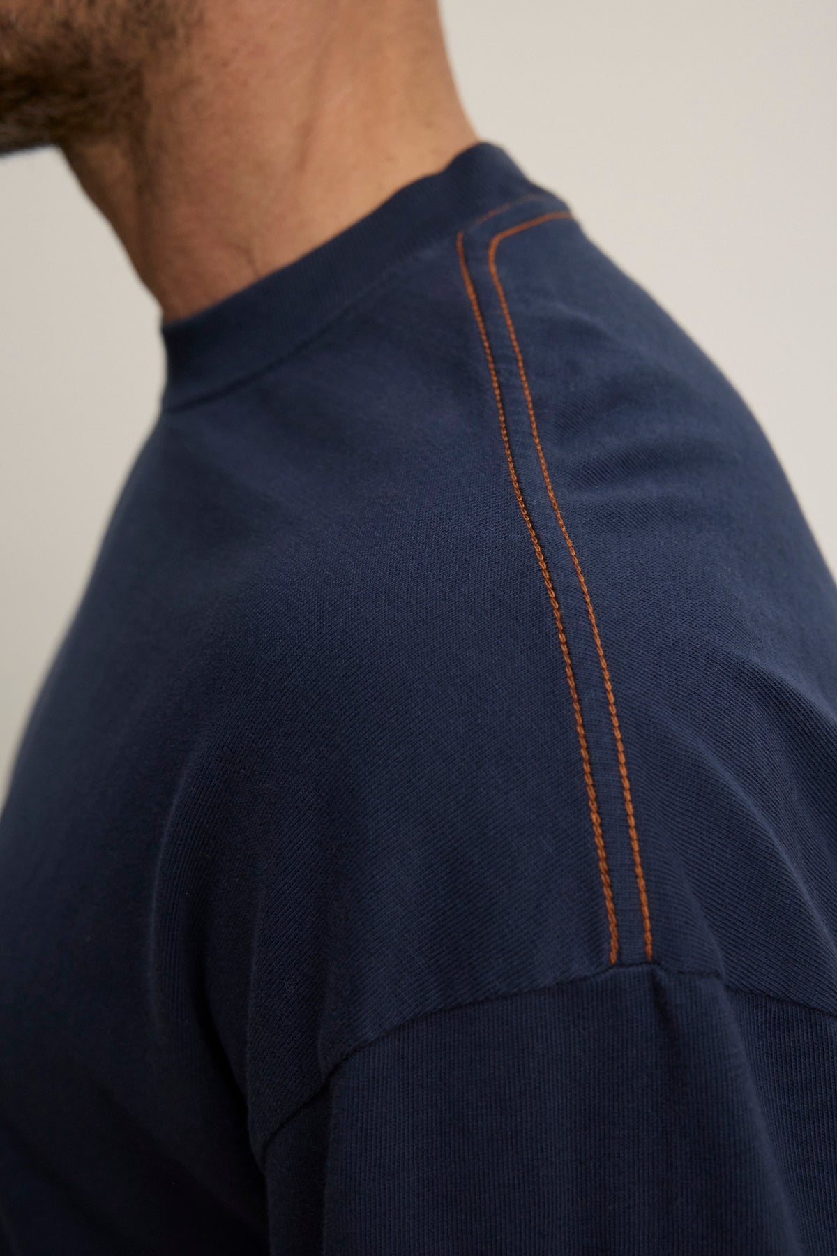   Close-up of someone wearing the SLATER TEE by Velvet by Graham & Spencer, a navy blue shirt made from soft pima cotton jersey, featuring a relaxed fit and orange contrast stitching on the shoulder seam. 