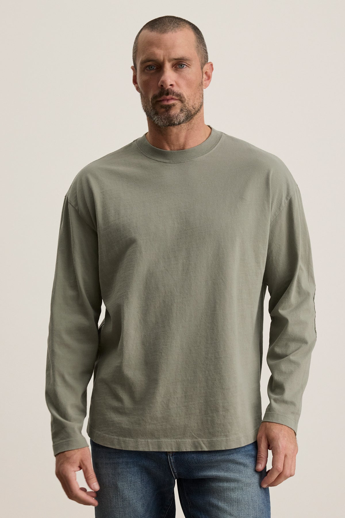   A bearded man with short hair wears the Velvet by Graham & Spencer SLATER TEE in light green, a relaxed-fit long-sleeve shirt made from structured Pima cotton, paired with blue jeans against a plain backdrop. 