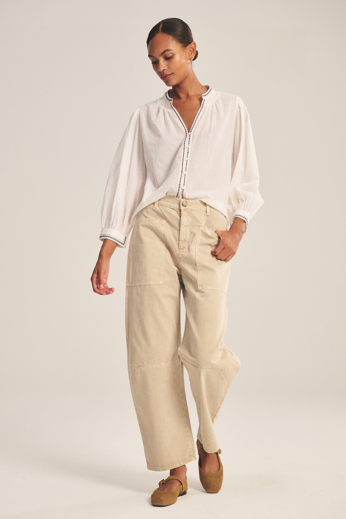 A woman stands against a plain background wearing a loose white blouse, brown flat shoes, and the BRYLIE PANT by Velvet by Graham & Spencer—cotton twill trousers with utilitarian patch pockets.