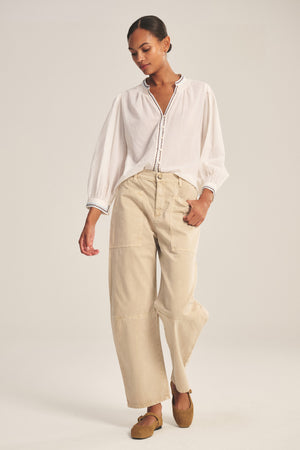 A woman stands against a plain background wearing a loose white blouse, brown flat shoes, and the BRYLIE PANT by Velvet by Graham & Spencer—cotton twill trousers with utilitarian patch pockets.