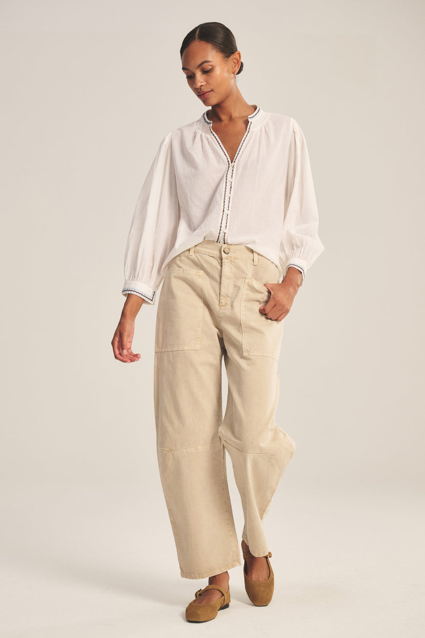 A woman stands against a plain background wearing a loose white blouse, brown flat shoes, and the BRYLIE PANT by Velvet by Graham & Spencer—cotton twill trousers with utilitarian patch pockets.