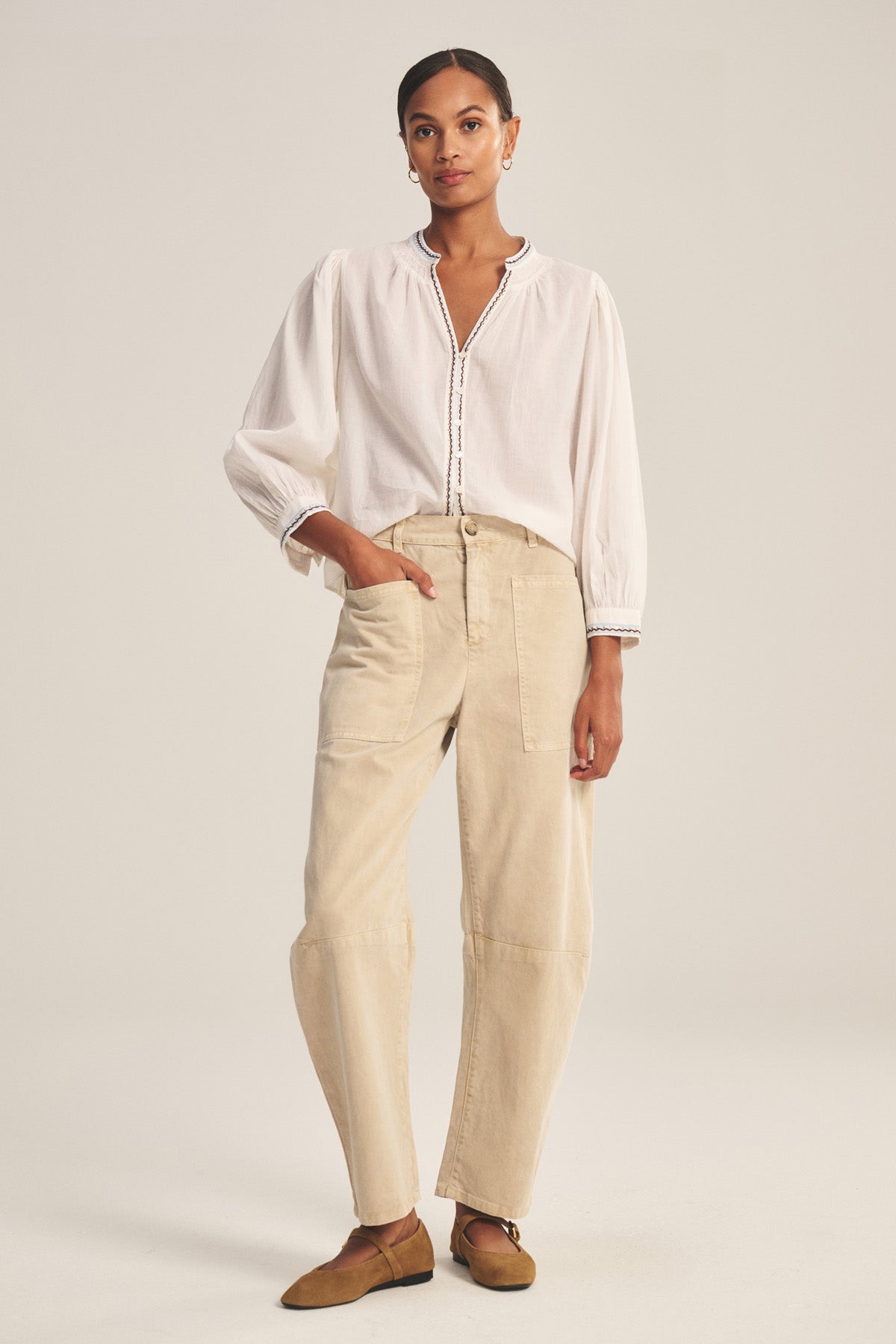 A woman stands in a neutral studio wearing the BRYLIE PANT by Velvet by Graham & Spencer, a beige cotton twill trouser with utilitarian patch pockets, paired with a white long-sleeve blouse and brown slip-on shoes.