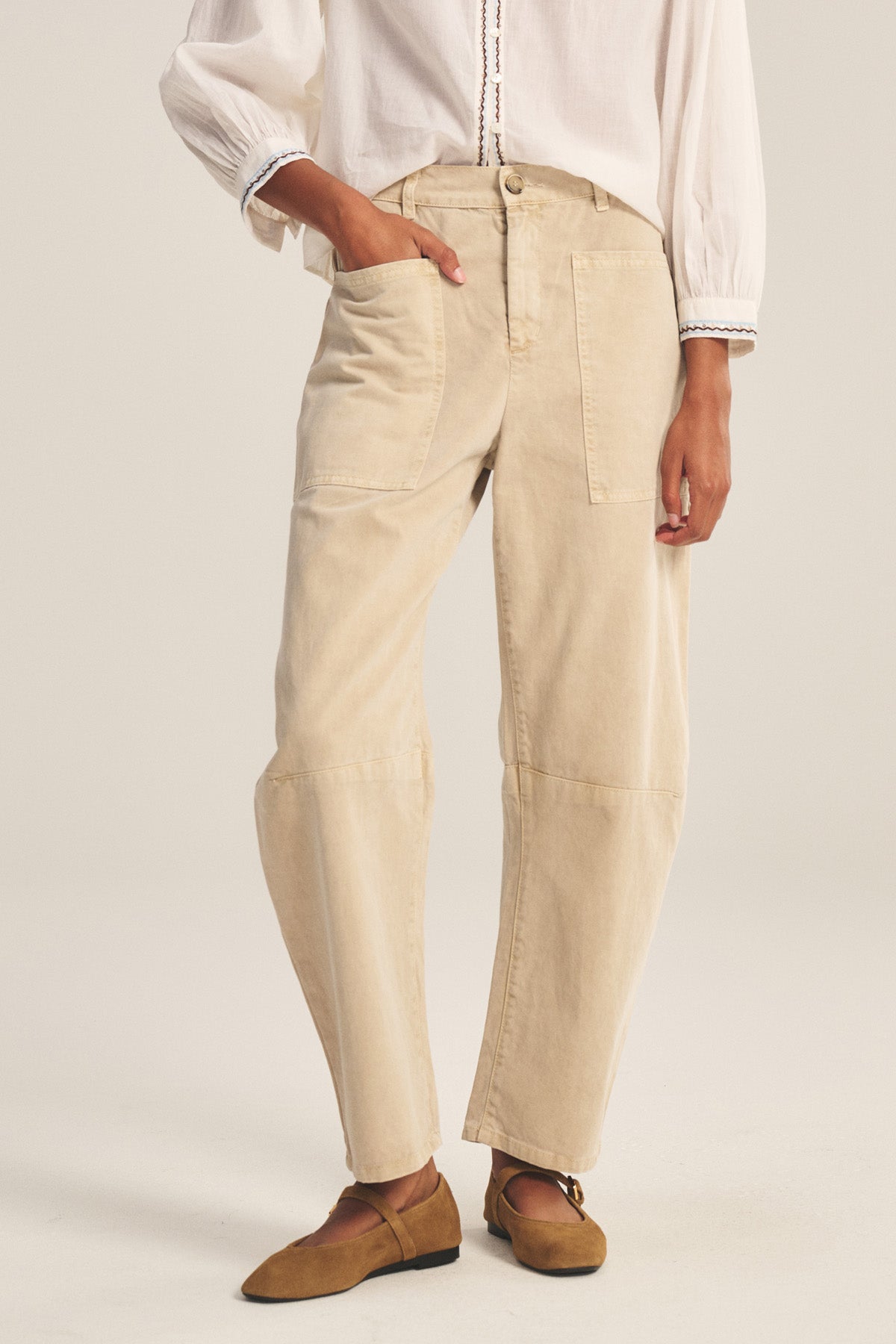 A person stands against a plain light background wearing a white long-sleeve blouse, tan flat shoes, and the BRYLIE PANT by Velvet by Graham & Spencer—a beige cotton twill trouser with utilitarian patch pockets.-41081200214209