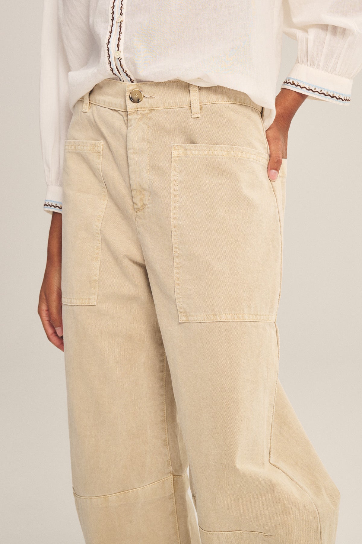 Model wears the BRYLIE PANT by Velvet by Graham & Spencer—beige cotton twill trousers with a curved silhouette and patch pockets—paired with a white long-sleeve blouse featuring black trim on cuffs and neckline.