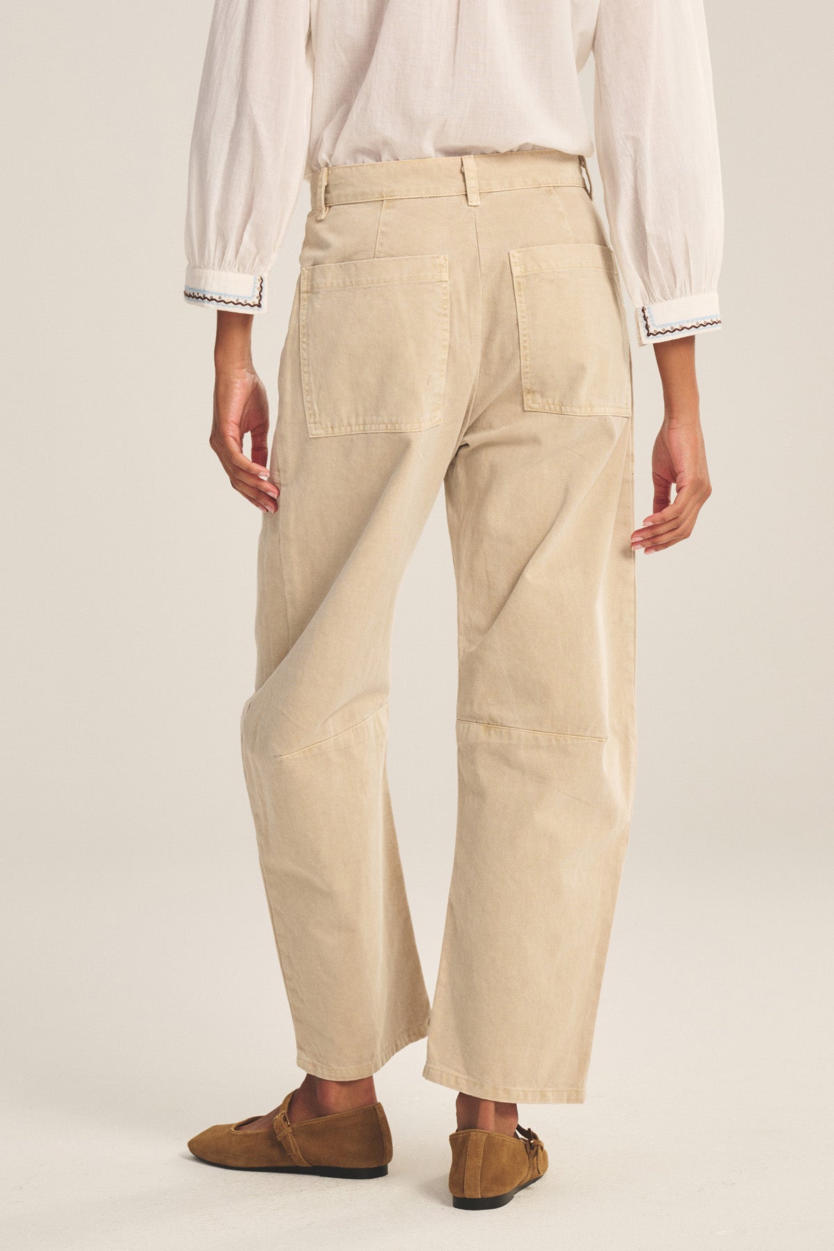 A person stands facing away, wearing a white long-sleeve shirt and Velvet by Graham & Spencer's BRYLIE PANT—beige cotton twill trousers with patch pockets and a curved silhouette—paired with brown flats against a plain background.