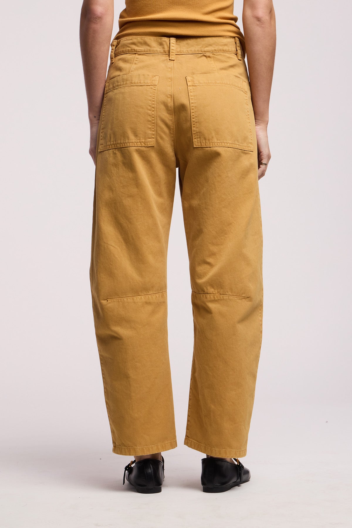 A person stands with their back to the camera against a plain background, wearing Velvet by Graham & Spencer’s BRYLIE PANT in mustard cotton twill with utilitarian patch pockets and black shoes.
