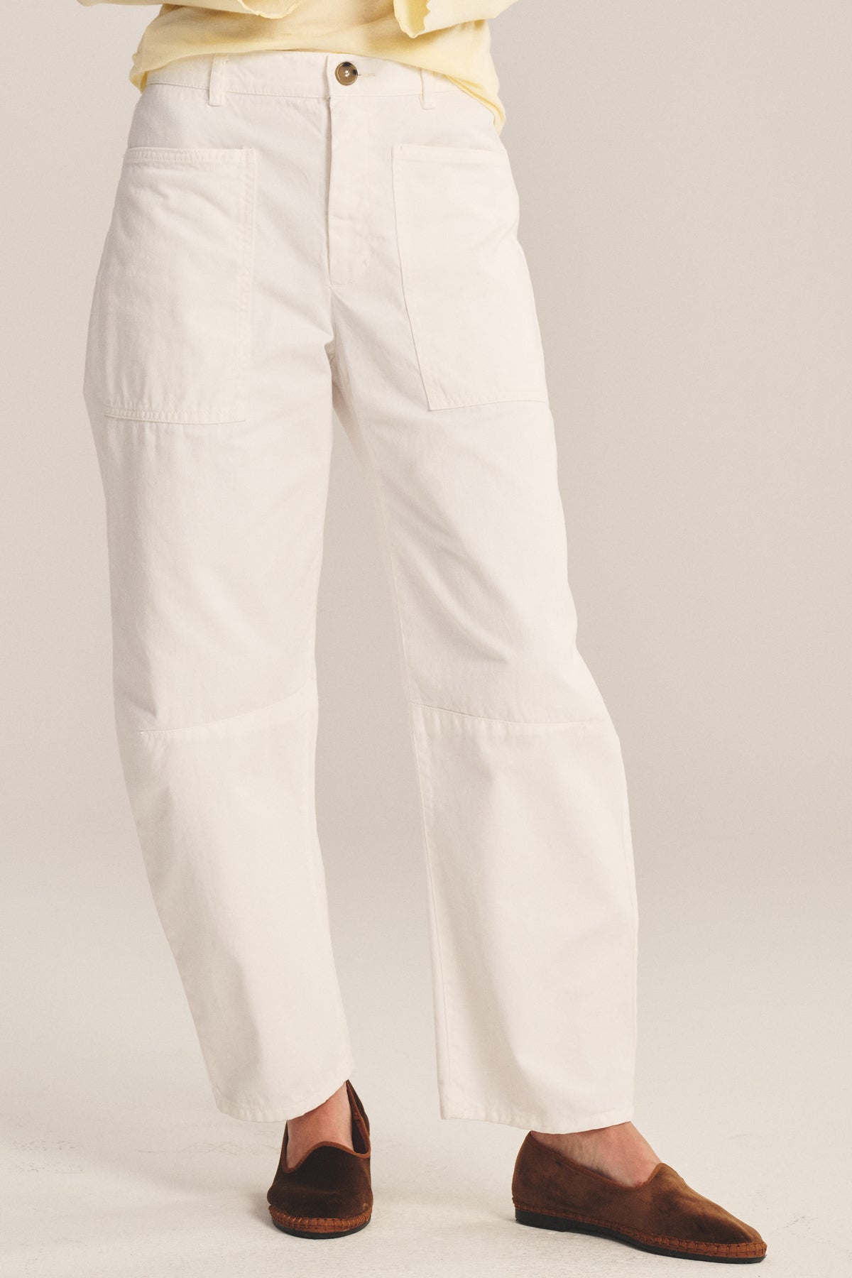 A person wears the BRYLIE PANT by Velvet by Graham & Spencer—white, straight-leg cotton twill trousers with utilitarian patch pockets—paired with brown slip-on shoes, standing against a neutral background.-41008452731073