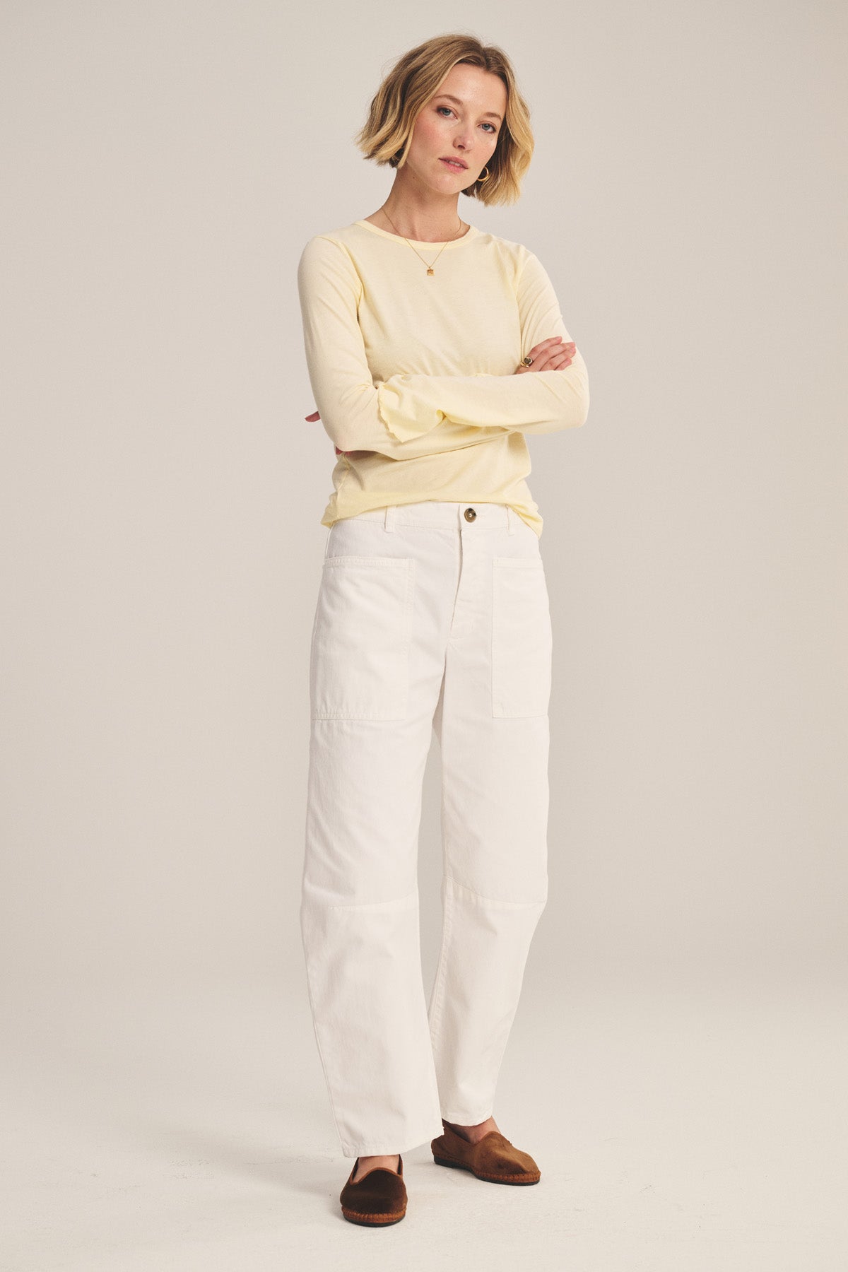 A woman stands with arms crossed, wearing a light yellow long-sleeve shirt with patch pockets, Velvet by Graham & Spencer's BRYLIE PANT in white cotton twill, and brown shoes against a plain background.-41008617357505