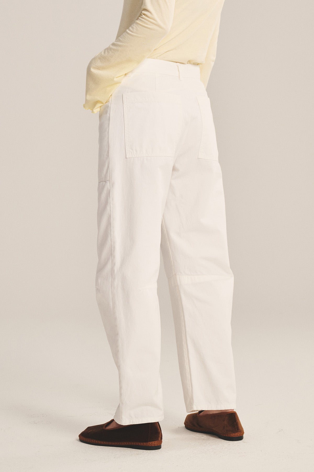 A person stands on a plain light background wearing Velvet by Graham & Spencer’s BRYLIE PANT, an off-white long-sleeve top, and brown flat shoes. The cotton twill trousers feature back and utilitarian patch pockets.