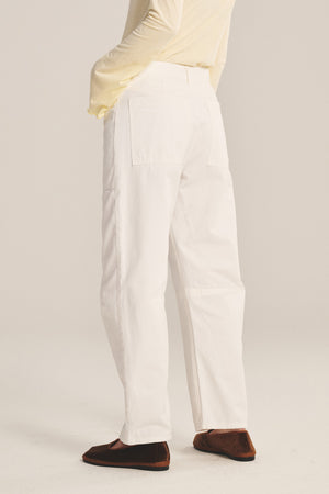 A person stands on a plain light background wearing Velvet by Graham & Spencer’s BRYLIE PANT, an off-white long-sleeve top, and brown flat shoes. The cotton twill trousers feature back and utilitarian patch pockets.