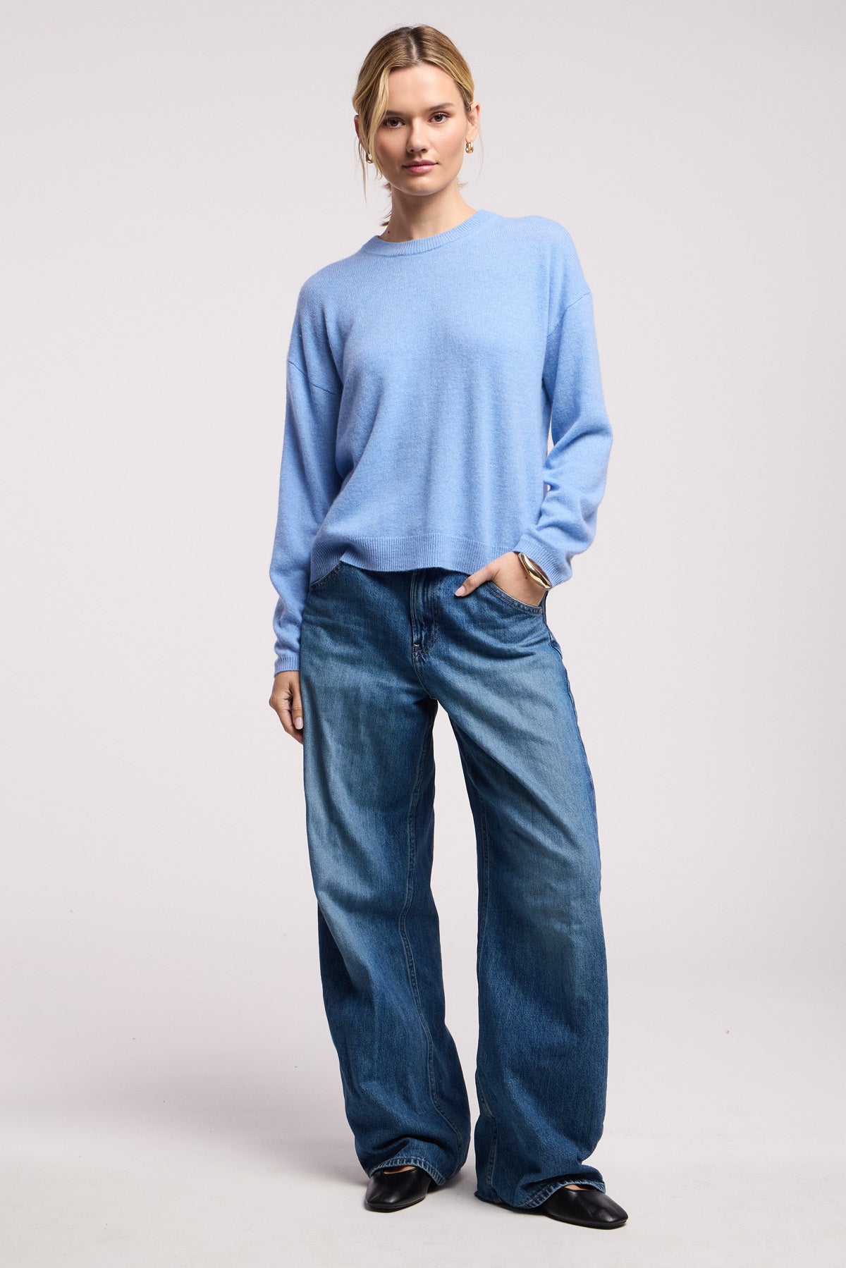 A person stands against a plain background wearing the BRYNNE CASHMERE SWEATER by Velvet by Graham & Spencer in light blue, paired with loose-fitting blue jeans and black shoes.-40521782853825