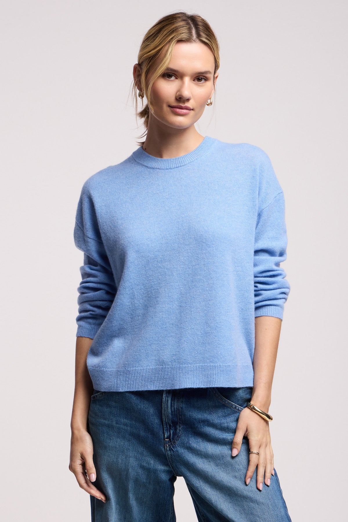 A woman wears the BRYNNE CASHMERE SWEATER by Velvet by Graham & Spencer—a light blue cashmere top with ribbed trims—paired with blue jeans, standing against a plain light background and looking at the camera neutrally.