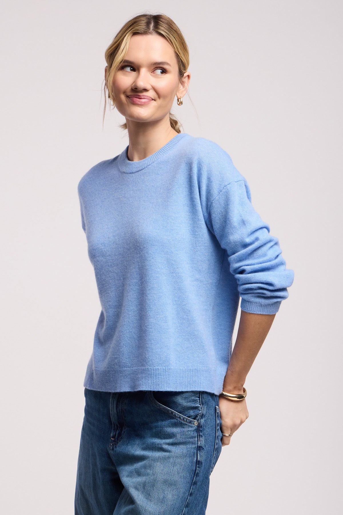 A woman wears the BRYNNE CASHMERE SWEATER by Velvet by Graham & Spencer in light blue with ribbed trims and blue jeans, standing against a plain background and looking to the side with a slight smile.-40521782558913